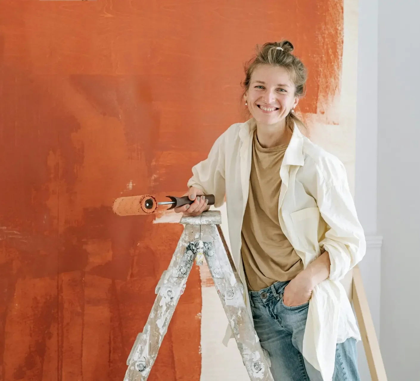 A cheerful woman in casual attire paints an orange wall while standing on a ladder indoors.